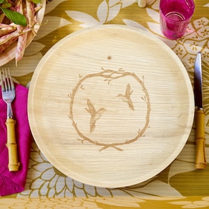 May include: A round, light-colored plate with a hummingbird design. The plate features a brown illustration of two hummingbirds within a circular branch frame. A star is above the frame. A fork, knife, and pink napkin are visible.