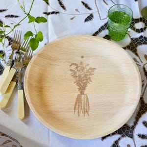 May include: A round, light-colored wooden plate with a detailed etched illustration of a carrot bouquet. The plate is surrounded by silverware with cream-colored handles and a green glass. The plate is approximately 10 inches in diameter.