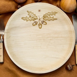 May include: A round, light beige palm leaf plate featuring a natural wood grain texture. The center of the plate is decorated with a brown line art illustration of three acorns attached to two oak leaves, topped by a small, simple sun symbol.