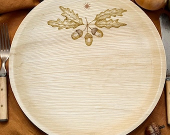 May include: A round, light beige palm leaf plate featuring a natural wood grain texture. The center of the plate is decorated with a brown line art illustration of three acorns attached to two oak leaves, topped by a small, simple sun symbol.