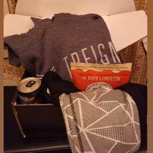 Puede incluir: Una caja de regalo abierta que contiene una sudadera gris con texto blanco, un paquete de pastel de prote&iacute;nas rojo y beige, una lata negra y una bufanda estampada.