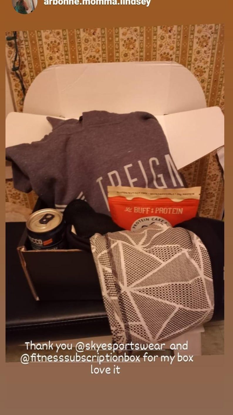 Puede incluir: Una caja de regalo abierta que contiene una sudadera gris con texto blanco, un paquete de pastel de prote&iacute;nas rojo y beige, una lata negra y una bufanda estampada.