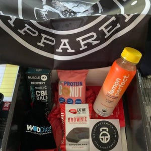 Puede incluir: Una caja llena de art&iacute;culos relacionados con el fitness. Incluye una botella de agua con sabor a lim&oacute;n, una rosquilla de prote&iacute;na, un brownie y una bolsa negra con el texto "WODDice". Se ve una pancarta con la palabra "APPAREL".