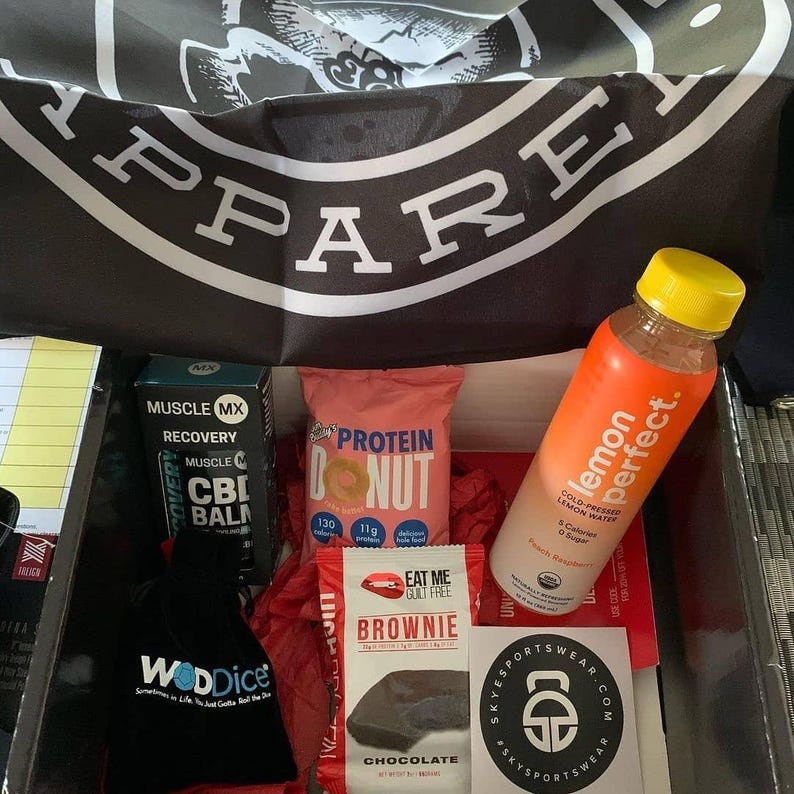 Puede incluir: Una caja llena de art&iacute;culos relacionados con el fitness. Incluye una botella de agua con sabor a lim&oacute;n, una rosquilla de prote&iacute;na, un brownie y una bolsa negra con el texto "WODDice". Se ve una pancarta con la palabra "APPAREL".