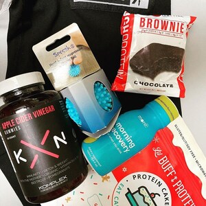 Puede incluir: Un surtido de productos de salud y bienestar, que incluyen un brownie, gomitas de vinagre de sidra de manzana, una bola de acupresi&oacute;n para manos y pies, mezcla para pastel de prote&iacute;nas y una botella de bebida azul. Sobre fondo negro.