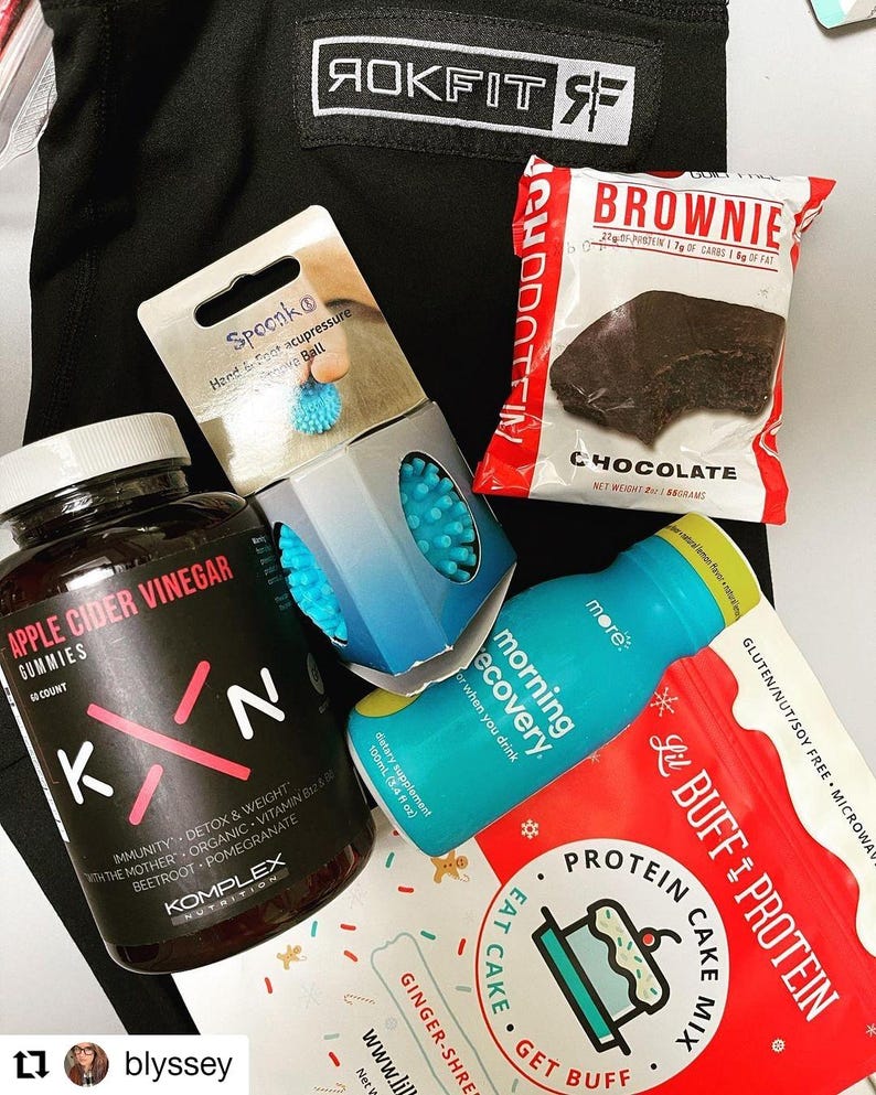 Puede incluir: Un surtido de productos de salud y bienestar, que incluyen un brownie, gomitas de vinagre de sidra de manzana, una bola de acupresi&oacute;n para manos y pies, mezcla para pastel de prote&iacute;nas y una botella de bebida azul. Sobre fondo negro.