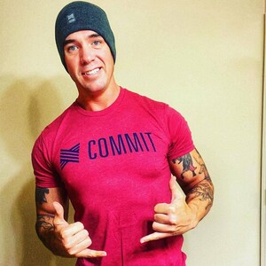 Puede incluir: Una camiseta roja con la palabra "COMMIT" en letras may&uacute;sculas azul marino. La camiseta se combina con un gorro gris oscuro. La persona est&aacute; haciendo una se&ntilde;al de aprobaci&oacute;n.