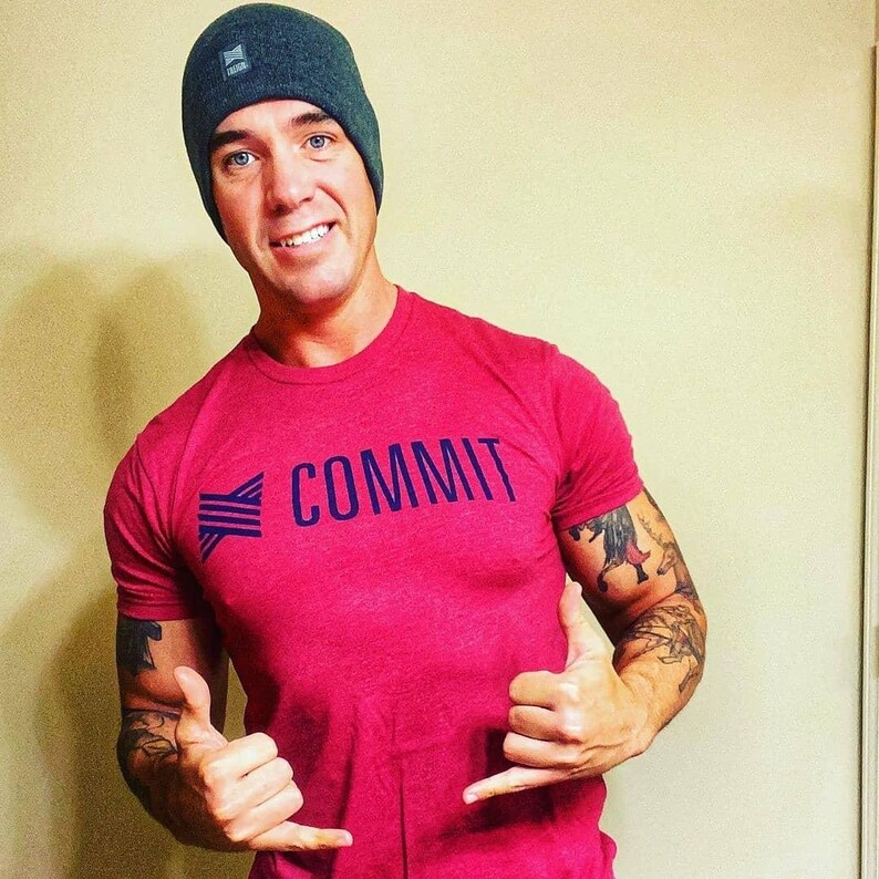 Puede incluir: Una camiseta roja con la palabra "COMMIT" en letras may&uacute;sculas azul marino. La camiseta se combina con un gorro gris oscuro. La persona est&aacute; haciendo una se&ntilde;al de aprobaci&oacute;n.