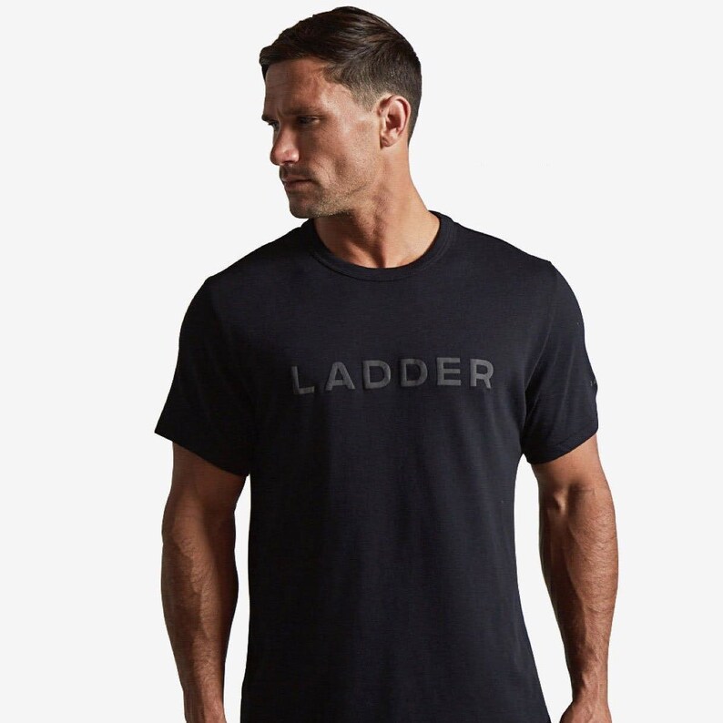 Puede incluir: Camiseta negra de cuello redondo con la palabra "LADDER" en relieve en el pecho. La camiseta tiene mangas cortas y un dise&ntilde;o minimalista. La tela parece ser un material suave y mate.