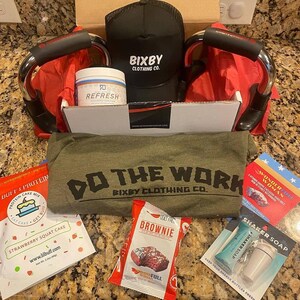Puede incluir: Una caja abierta llena de productos de fitness y bienestar. Incluye una gorra negra con el texto "Bixby Clothing Co.", un tarro con la etiqueta "Refresh", barras para flexiones, una camiseta verde con el texto "Do The Work" y varios paquetes de alimentos.