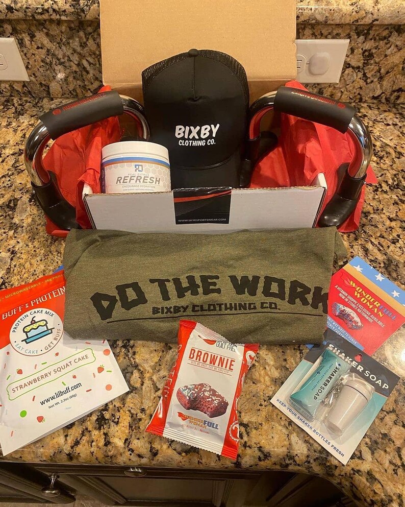 Puede incluir: Una caja abierta llena de productos de fitness y bienestar. Incluye una gorra negra con el texto "Bixby Clothing Co.", un tarro con la etiqueta "Refresh", barras para flexiones, una camiseta verde con el texto "Do The Work" y varios paquetes de alimentos.
