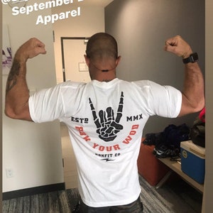 Puede incluir: Camiseta blanca con un gr&aacute;fico de mano de esqueleto negro que dice "Rock Your WOD" y "Rokfit Co.". La camiseta tambi&eacute;n tiene el texto "ESTO MMX". La persona lleva pantalones cortos de camuflaje.