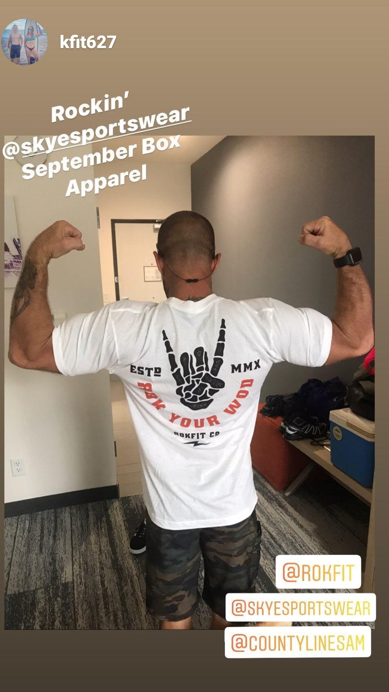 Puede incluir: Camiseta blanca con un gr&aacute;fico de mano de esqueleto negro que dice "Rock Your WOD" y "Rokfit Co.". La camiseta tambi&eacute;n tiene el texto "ESTO MMX". La persona lleva pantalones cortos de camuflaje.