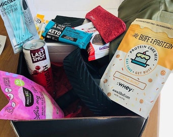 Op de afbeelding: Een zwarte geschenkdoos gevuld met diverse items, waaronder een blikje Last Shot kersendrank, Miracle Rice, eiwitrepen, een rode sok en een Lil Buff Protein cakemix. Ook inbegrepen zijn Hickies vetersysteem en groene leggings.