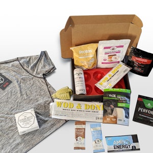 Puede incluir: Una camiseta deportiva gris con ribetes negros y una etiqueta, junto a una caja de cart&oacute;n llena de varios suplementos de fitness y mezclas de prote&iacute;nas. La caja tambi&eacute;n contiene un molde de silicona rojo para donuts y una botella de Fit Bag.
