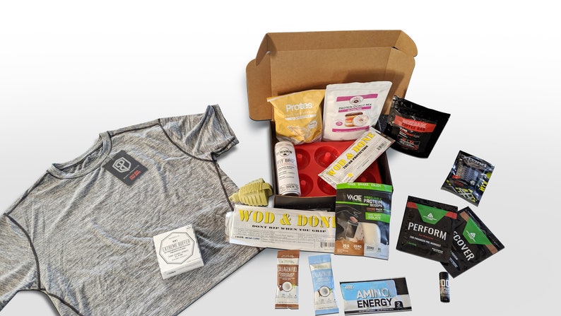 Puede incluir: Una camiseta deportiva gris con ribetes negros y una etiqueta, junto a una caja de cart&oacute;n llena de varios suplementos de fitness y mezclas de prote&iacute;nas. La caja tambi&eacute;n contiene un molde de silicona rojo para donuts y una botella de Fit Bag.