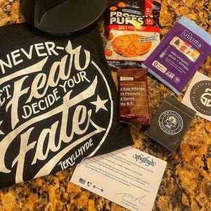 Puede incluir: Un conjunto de art&iacute;culos de fitness y nutrici&oacute;n, que incluye una camiseta negra con el texto "NEVER FEAR DECIDE YOUR FATE" y un asa negra. Tambi&eacute;n se incluyen puffs de prote&iacute;nas, una barrita de prote&iacute;nas y sobres de electrolitos.