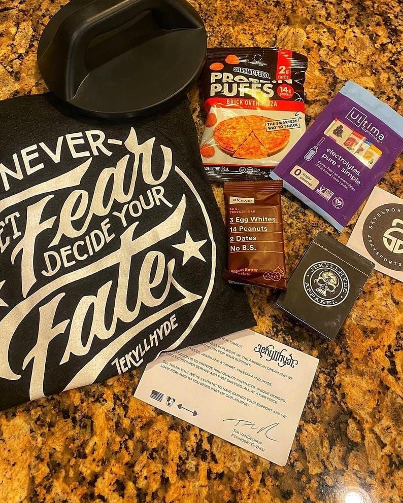 Puede incluir: Un conjunto de art&iacute;culos de fitness y nutrici&oacute;n, que incluye una camiseta negra con el texto "NEVER FEAR DECIDE YOUR FATE" y un asa negra. Tambi&eacute;n se incluyen puffs de prote&iacute;nas, una barrita de prote&iacute;nas y sobres de electrolitos.