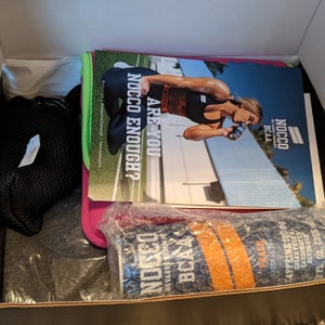 Puede incluir: Una caja que contiene productos de fitness y salud. Incluye una revista con el texto "ARE YOU NOCCO ENOUGH?" y una lata de bebida NOCCO BCAA con sabor a melocot&oacute;n. Tambi&eacute;n son visibles una bolsa de malla negra, una camiseta gris y un art&iacute;culo rosa y verde.