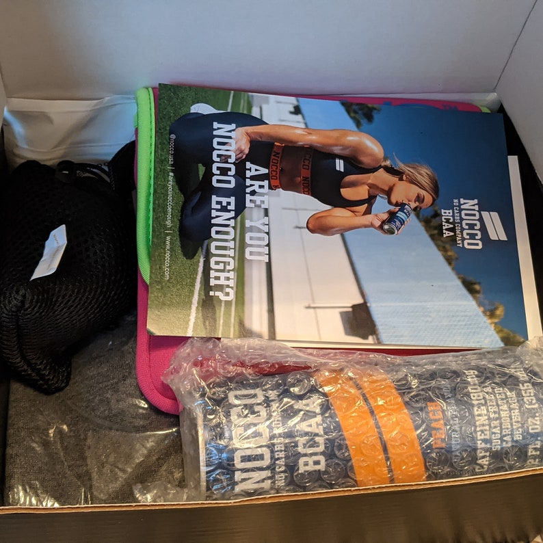 Puede incluir: Una caja que contiene productos de fitness y salud. Incluye una revista con el texto "ARE YOU NOCCO ENOUGH?" y una lata de bebida NOCCO BCAA con sabor a melocot&oacute;n. Tambi&eacute;n son visibles una bolsa de malla negra, una camiseta gris y un art&iacute;culo rosa y verde.