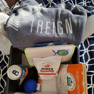 Puede incluir: Una caja abierta que contiene varios art&iacute;culos. Se ve una sudadera con capucha gris con la palabra "TREIGN" en blanco. Otros art&iacute;culos incluyen una funda "yogo" blanca y verde, una lata de bebida "NOCCo", un paquete de sales minerales "Mike's Recovery", una bola azul y una mezcla para pastel "Lil Buff & Protein".