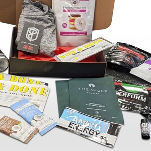 Puede incluir: Una caja abierta llena de productos de fitness y bienestar. Incluye una camiseta deportiva gris, mezcla de prote&iacute;nas, bebida energ&eacute;tica, mascarilla facial, barritas de prote&iacute;nas y varios paquetes de suplementos. La caja es negra con interior rojo.