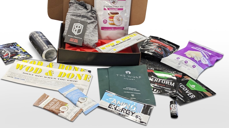 Puede incluir: Una caja abierta llena de productos de fitness y bienestar. Incluye una camiseta deportiva gris, mezcla de prote&iacute;nas, bebida energ&eacute;tica, mascarilla facial, barritas de prote&iacute;nas y varios paquetes de suplementos. La caja es negra con interior rojo.