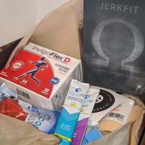 Puede incluir: Una caja llena de productos de salud y fitness. Los art&iacute;culos incluyen una caja de suplemento para las articulaciones InvigoFlex D, sobres de mezcla de hidrataci&oacute;n Liquid I.V. y una cuerda de velocidad JerkFit Omega. La caja est&aacute; forrada con papel marr&oacute;n.