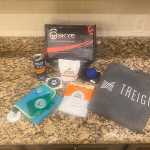 Puede incluir: Una caja de fitness con varios art&iacute;culos, incluyendo una caja negra con el texto "SKYE SPORTSWEAR", una lata de NOCCO BCAA, una bolsa de Mike's Recovery, una bola de masaje azul, una bolsa de mano gris con el texto "TREIGO" y otros productos de fitness.