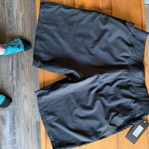 Puede incluir: Pantalones cortos deportivos negros con una cintura ancha y una etiqueta adjunta. Los pantalones cortos est&aacute;n hechos de un material ligero. Los pantalones cortos est&aacute;n sobre una superficie de madera.
