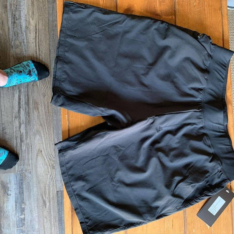 Puede incluir: Pantalones cortos deportivos negros con una cintura ancha y una etiqueta adjunta. Los pantalones cortos est&aacute;n hechos de un material ligero. Los pantalones cortos est&aacute;n sobre una superficie de madera.