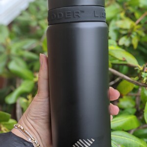 Puede incluir: Una botella de agua aislada negra con la palabra "LADDER" impresa en blanco. La botella se sostiene en una mano y tiene una tapa negra y una banda negra con la palabra "LADDER" en relieve.