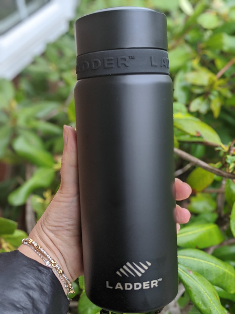 Puede incluir: Una botella de agua aislada negra con la palabra "LADDER" impresa en blanco. La botella se sostiene en una mano y tiene una tapa negra y una banda negra con la palabra "LADDER" en relieve.