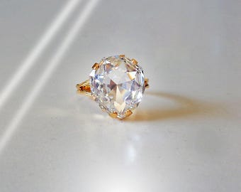 May include: A gold ring featuring a large, clear, pear-shaped faceted gemstone held in place by multiple prongs. The band is thin and metallic, showcasing a reflective surface that catches the light, creating a bright, sparkling appearance.