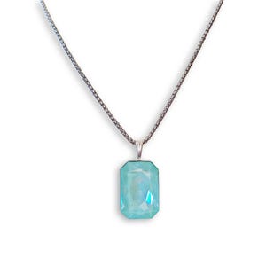 Puede incluir: Un collar de cadena plateada con un colgante rectangular azul claro. El colgante tiene una apariencia facetada, similar a una gema, que captura la luz. El collar se muestra sobre un fondo blanco, resaltando el diseño de la joya.