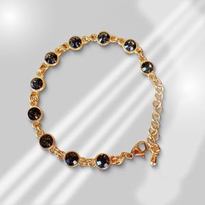 Puede incluir: Un brazalete de cadena dorada con piedras redondas de color gris oscuro. El brazalete tiene un cierre de langosta.