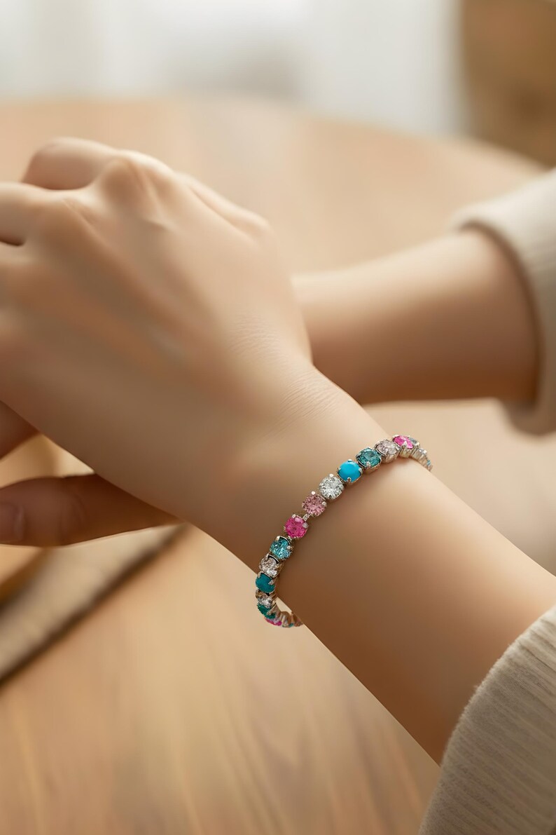 May include: A colorful bracelet with round gemstones in shades of turquoise, pink, and clear white. The bracelet is worn on a wrist, with the gemstones set in a silver-colored metal. The bracelet is a piece of jewelry.