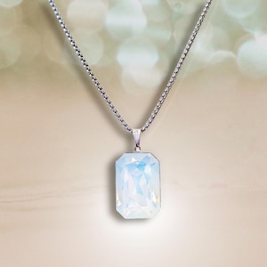 Peut inclure: Un collier argenté avec un pendentif rectangulaire bleu clair. Le pendentif présente un design à facettes, et la chaîne est de style délicat et maillé. Le fond est un dégradé flou de beige et de blanc.
