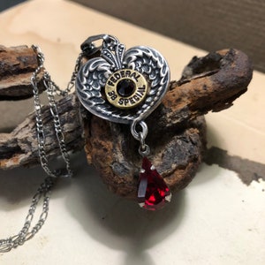 May include: Silver heart pendant necklace with a bullet casing featuring the text "FEDERAL 38 SPECIAL" and a red gemstone. The pendant is attached to a silver chain.