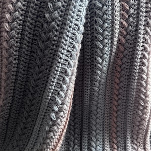 May include: A close-up of a handmade crochet blanket in shades of gray and beige. The blanket features a textured pattern with vertical and diagonal stitch designs. The soft, folded fabric creates shadows and highlights, showcasing the intricate craftsmanship.