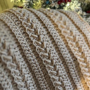 K&ouml;nnte beinhalten: Nahaufnahme einer beige geh&auml;kelten Decke mit einem strukturierten, geflochtenen Design. Die Decke weist vertikale Rippenabschnitte auf, die sich mit detaillierten Webmustern abwechseln. Die weiche, neutrale Farbe und die komplizierte Sticharbeit deuten auf einen handgefertigten, gem&uuml;tlichen Artikel hin.