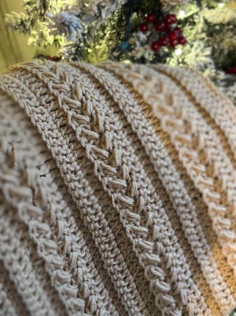 K&ouml;nnte beinhalten: Nahaufnahme einer beige geh&auml;kelten Decke mit einem strukturierten, geflochtenen Design. Die Decke weist vertikale Rippenabschnitte auf, die sich mit detaillierten Webmustern abwechseln. Die weiche, neutrale Farbe und die komplizierte Sticharbeit deuten auf einen handgefertigten, gem&uuml;tlichen Artikel hin.