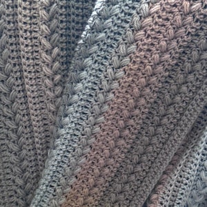 May include: A close-up shot of a grey crocheted blanket with a textured pattern. The blanket has a combination of raised stitches and openwork, creating a visually interesting surface. The colour is a muted grey, with subtle variations in tone.
