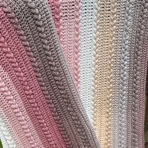 Puede incluir: Primer plano de una manta de ganchillo hecha a mano en un degradado de colores, incluyendo tonos de rosa, blanco, beige y marrón. La manta presenta un diseño texturizado con filas verticales de patrones trenzados.