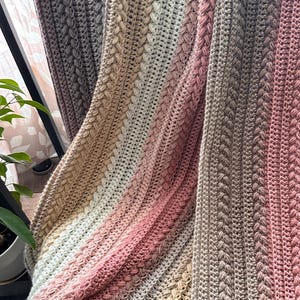 Op de afbeelding: Een handgehaakte deken in een kleurengradatie van lichtbeige tot zachtroze en grijs. De deken heeft een textuur kabelsteekontwerp, waardoor een gezellig en decoratief item ontstaat. De deken is over een stoel gedrapeerd.