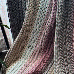 May include: A hand-knitted blanket in a mix of grey, green, and pink hues. The blanket has a detailed, woven design, offering a tactile and attractive texture. The colours merge, producing a gradient effect, perfect for home decor.