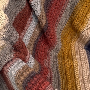 May include: Close-up of a handmade, textured blanket with a variety of colors. The blanket features stripes of gray, brown, red, and yellow. The knitted or crocheted design creates a cozy, warm appearance.