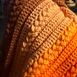 K&ouml;nnte beinhalten: Eine handgeh&auml;kelte Decke in Orange-, Pfirsich- und Braunt&ouml;nen. Die Decke hat ein strukturiertes Muster mit erhabenen Maschen, das ein gem&uuml;tliches und warmes Aussehen erzeugt. Der Farbverlauf verleiht dem handgefertigten Artikel visuelles Interesse.