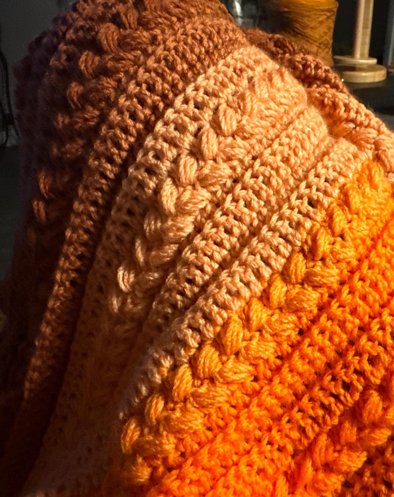 K&ouml;nnte beinhalten: Eine handgeh&auml;kelte Decke in Orange-, Pfirsich- und Braunt&ouml;nen. Die Decke hat ein strukturiertes Muster mit erhabenen Maschen, das ein gem&uuml;tliches und warmes Aussehen erzeugt. Der Farbverlauf verleiht dem handgefertigten Artikel visuelles Interesse.