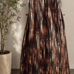 May include: A crochet blanket featuring a vertical striped pattern in shades of black, dark brown, copper, and cream. The textile has a textured, ribbed appearance created by repeating rows of yarn stitches, hanging draped over a wooden ladder frame.